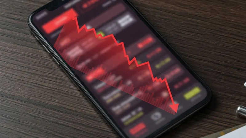 A smartphone on a wooden desk displays a declining red stock market graph with a downward arrow overlay.